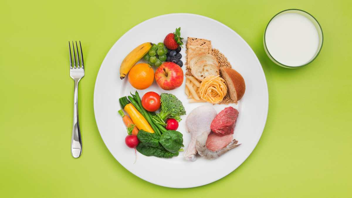Prato branco dividido em porções de legumes, carne e massa, ao lado de um copo de leite